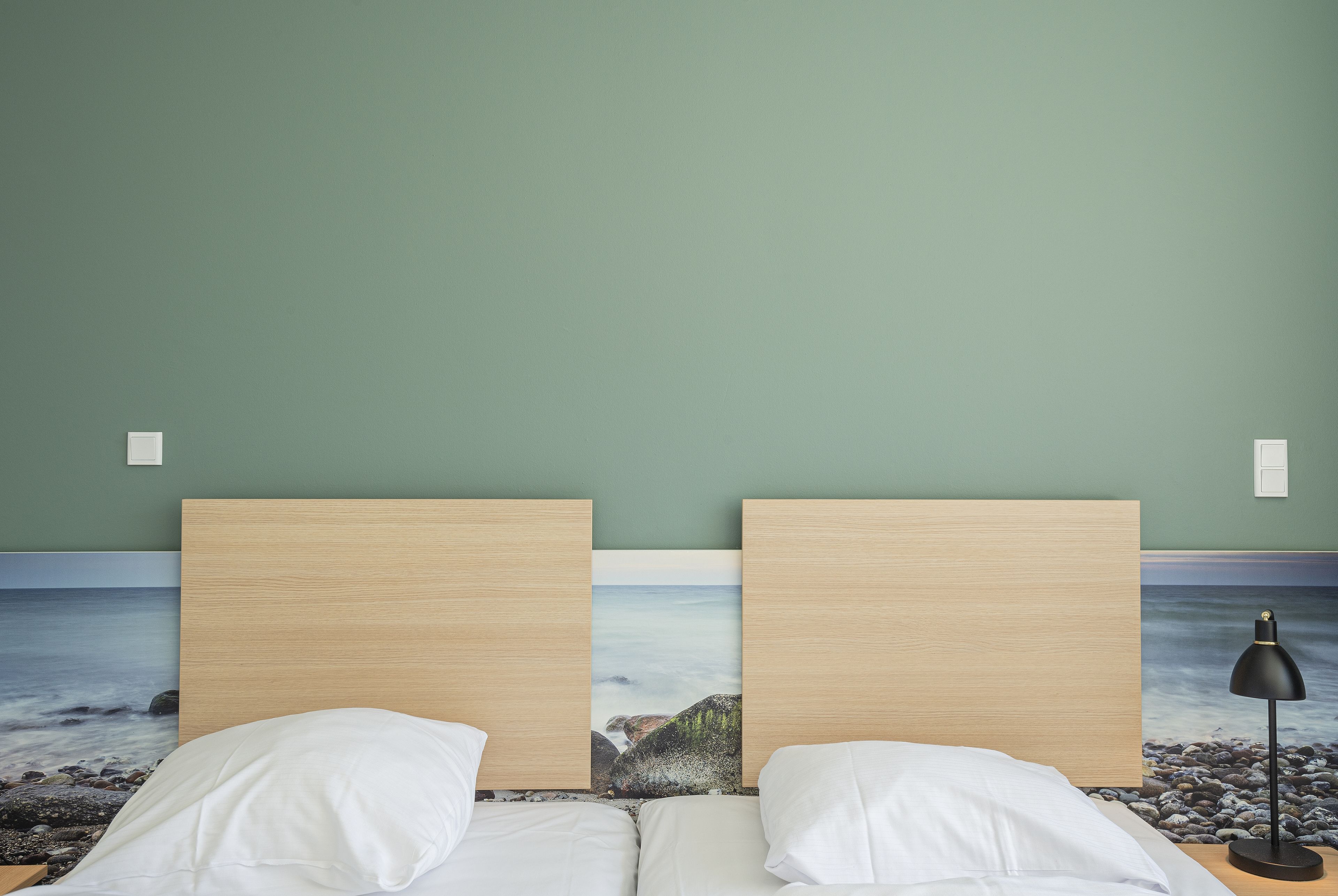 A double bed with white linens, a nightstand with a black lamp, and a wall mural of a beach scene.