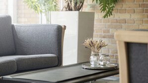 Una sala moderna con un sofá gris, una mesita con un jarrón de flores secas y una planta grande en maceta al fondo.