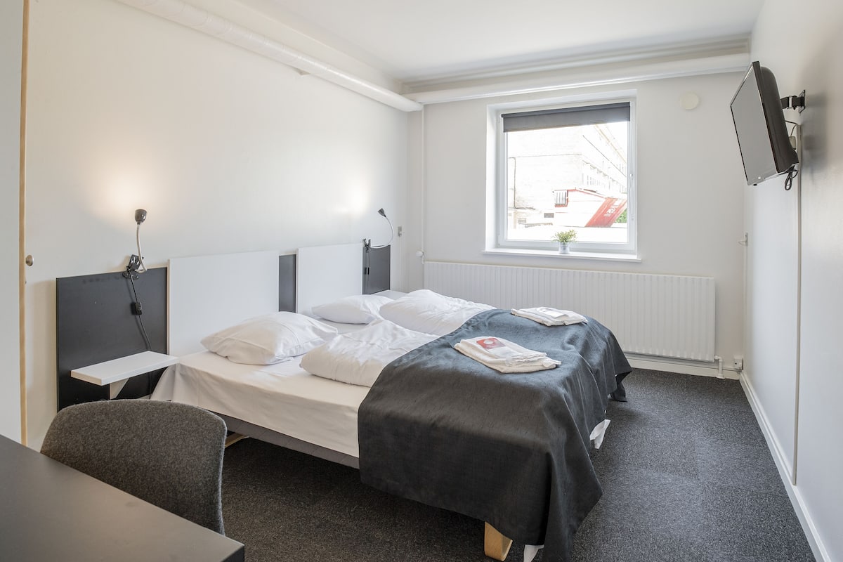 Une chambre d’hôtel de taille modeste, comprenant un lit simple, un bureau et une fenêtre donnant sur les bâtiments environnants.