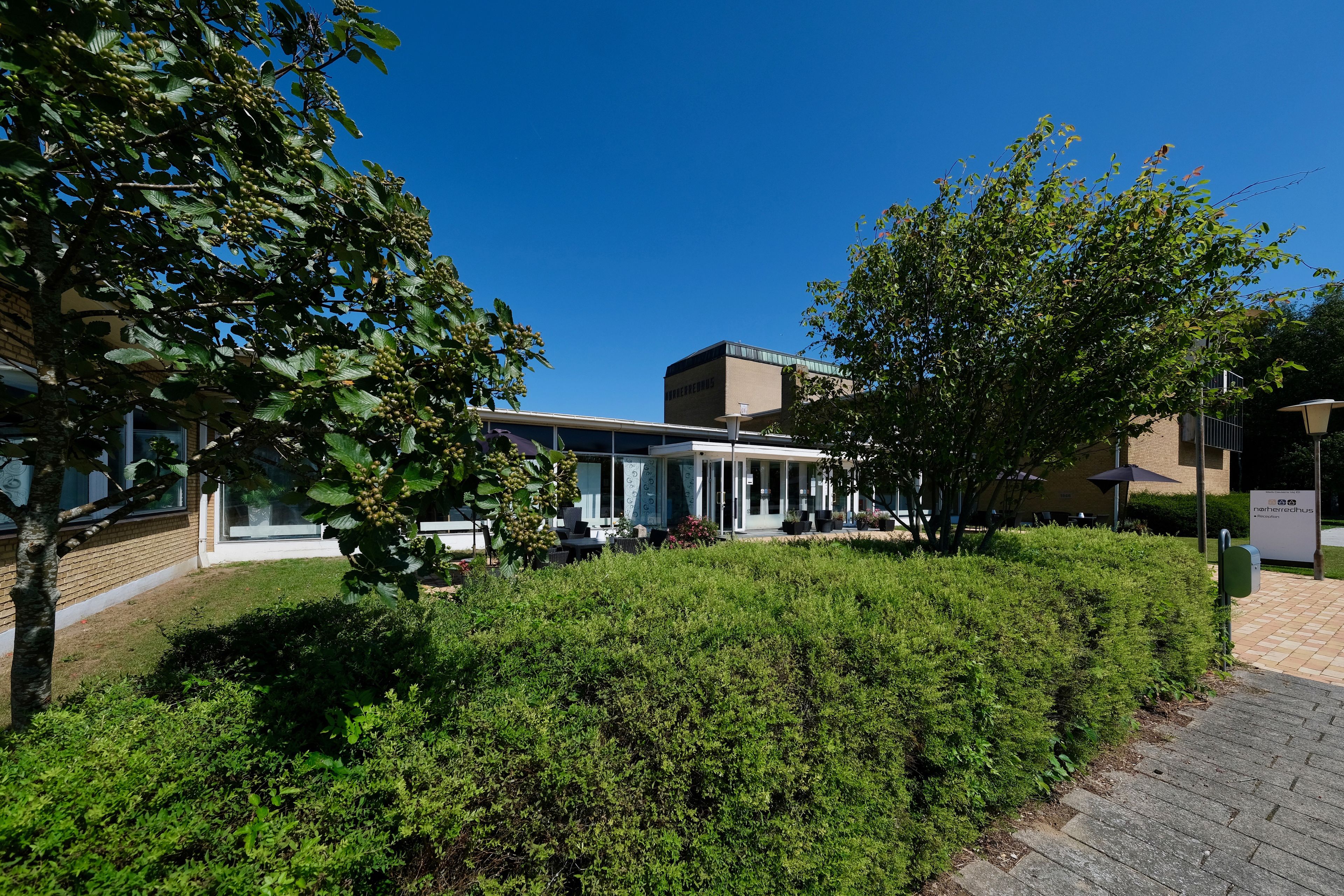Een modern gebouw met een glazen gevel, omgeven door goed onderhouden hagen en bomen.