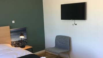 Habitación de hotel con cama, sillón, escritorio con lámpara, televisor de pantalla plana en la pared y un cuadro.