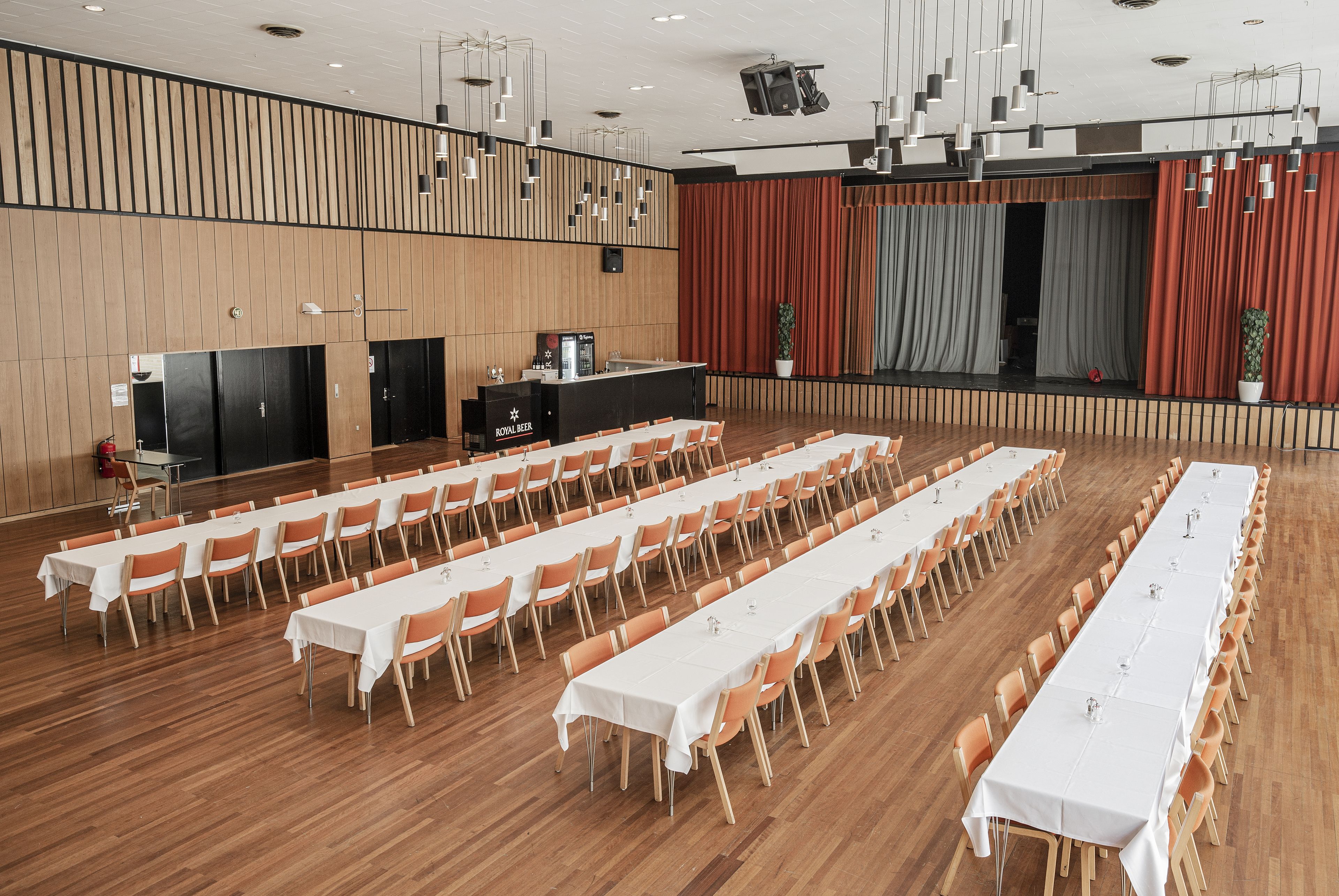 Ein großer Konferenzraum mit langen Tischen, Stühlen, einem Rednerpult und einer Bühne mit Vorhängen.
