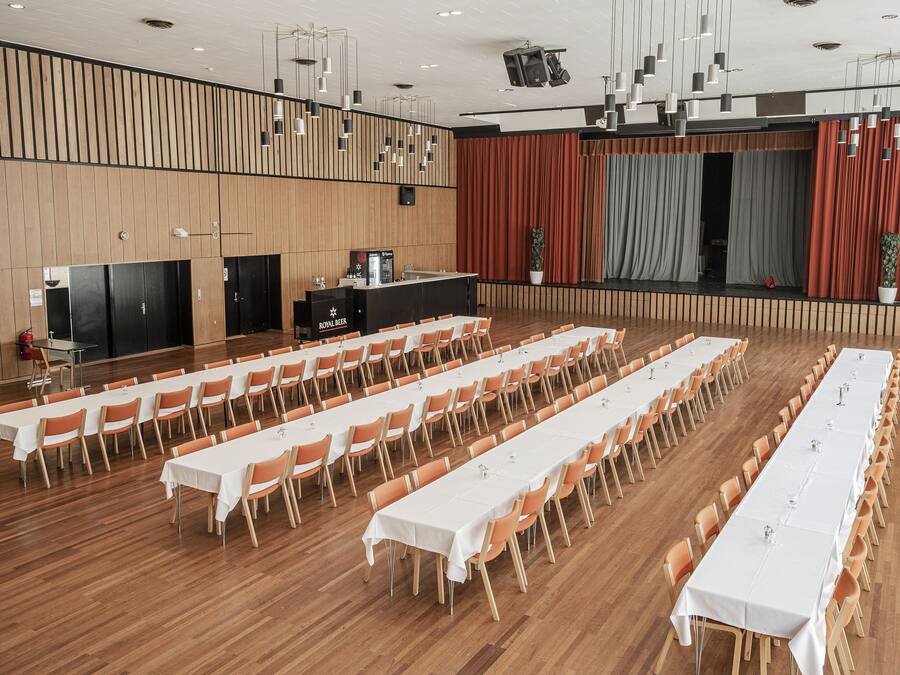 Un amplio salón de conferencias con mesas largas y sillas, un atril y un escenario con cortinas.