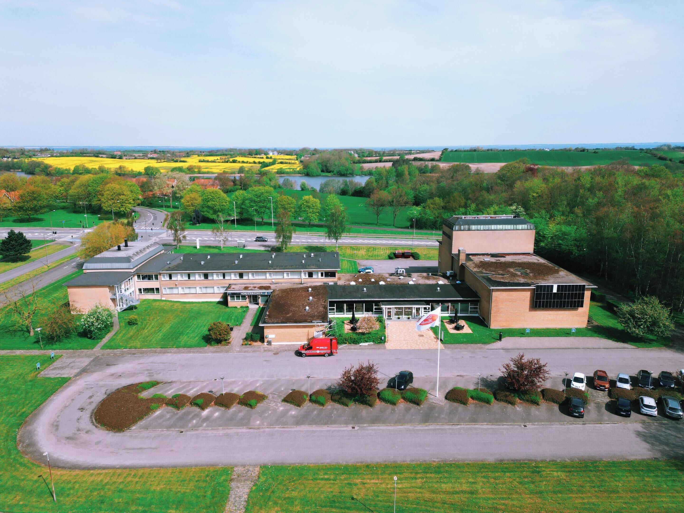 An aerial view of a building complex with multiple parking areas, a flag, and surrounding green fields.
