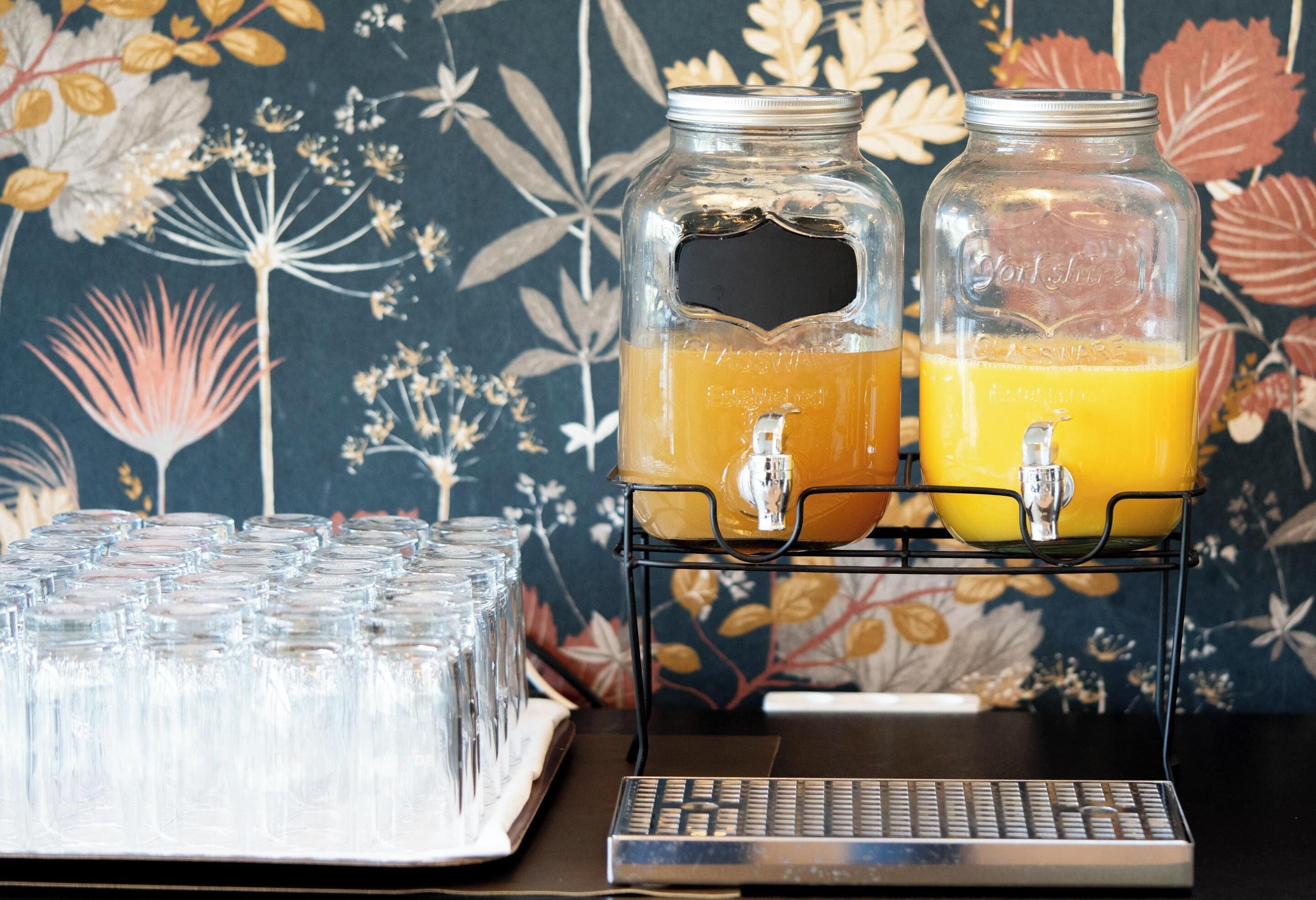 Una estación de bebidas con dos dispensadores de vidrio llenos de jugo de naranja, una bandeja de vasos vacíos y un teclado sobre una mesa.