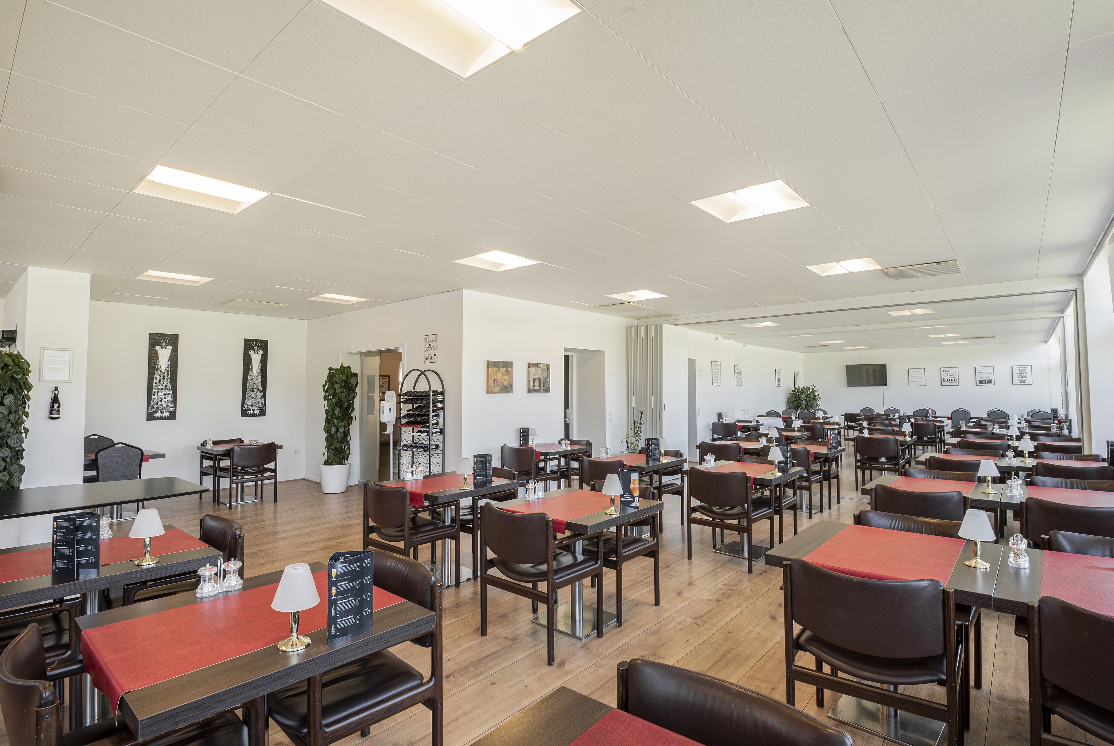 Een ruime eetruimte met meerdere tafels en stoelen, een houten vloer en plafondlampen.