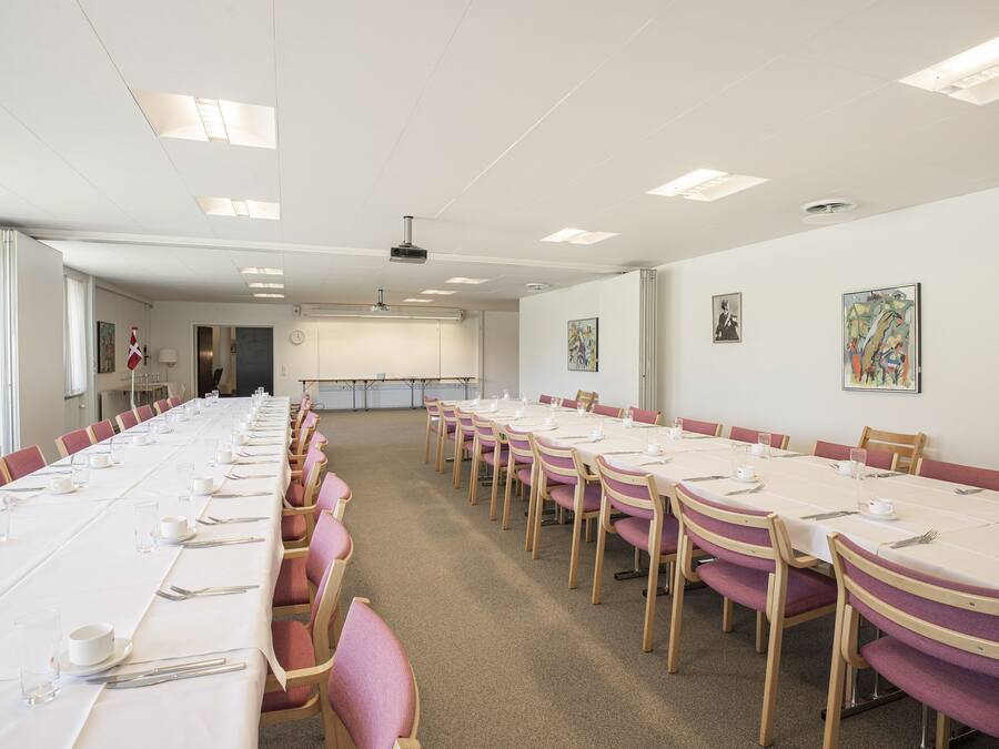 A conference room with long tables set for meetings, equipped with chairs and tables, and a projector on the ceiling.