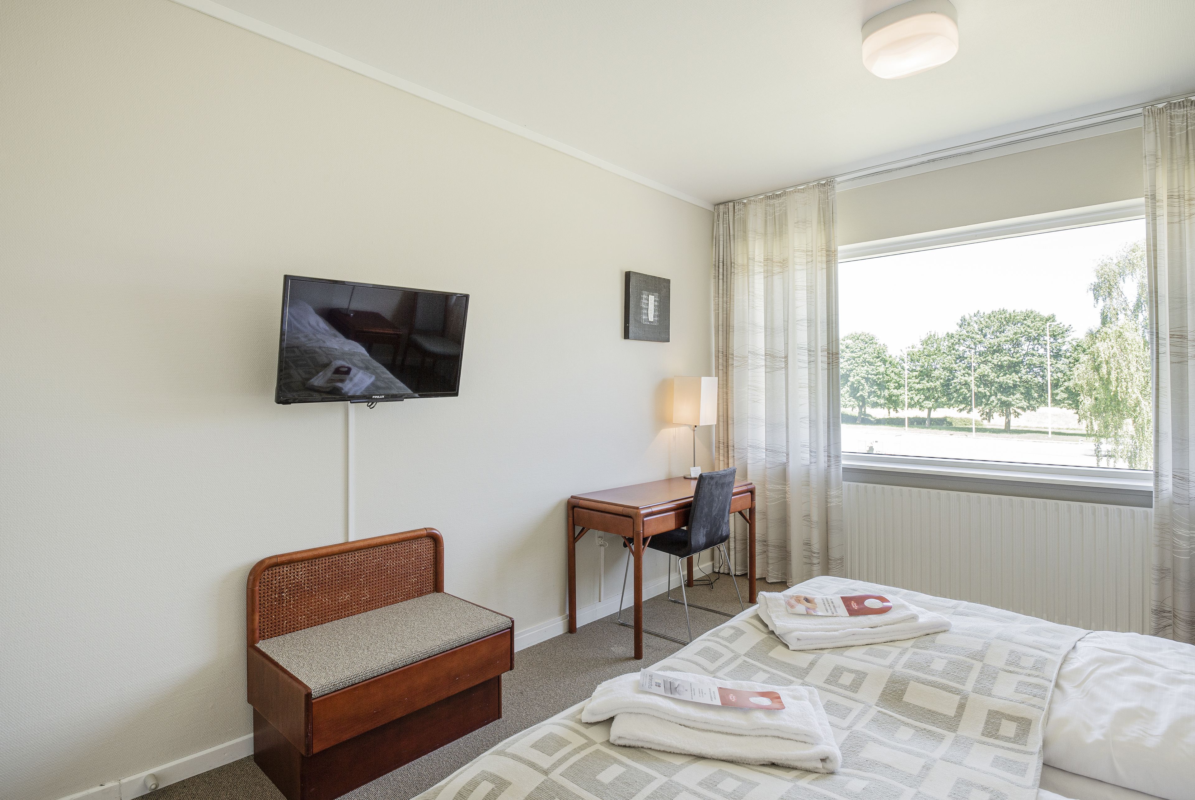 Une chambre d’hôtel équipée d’un lit, d’un bureau, d’une chaise, d’une télévision et d’une fenêtre donnant sur la verdure.
