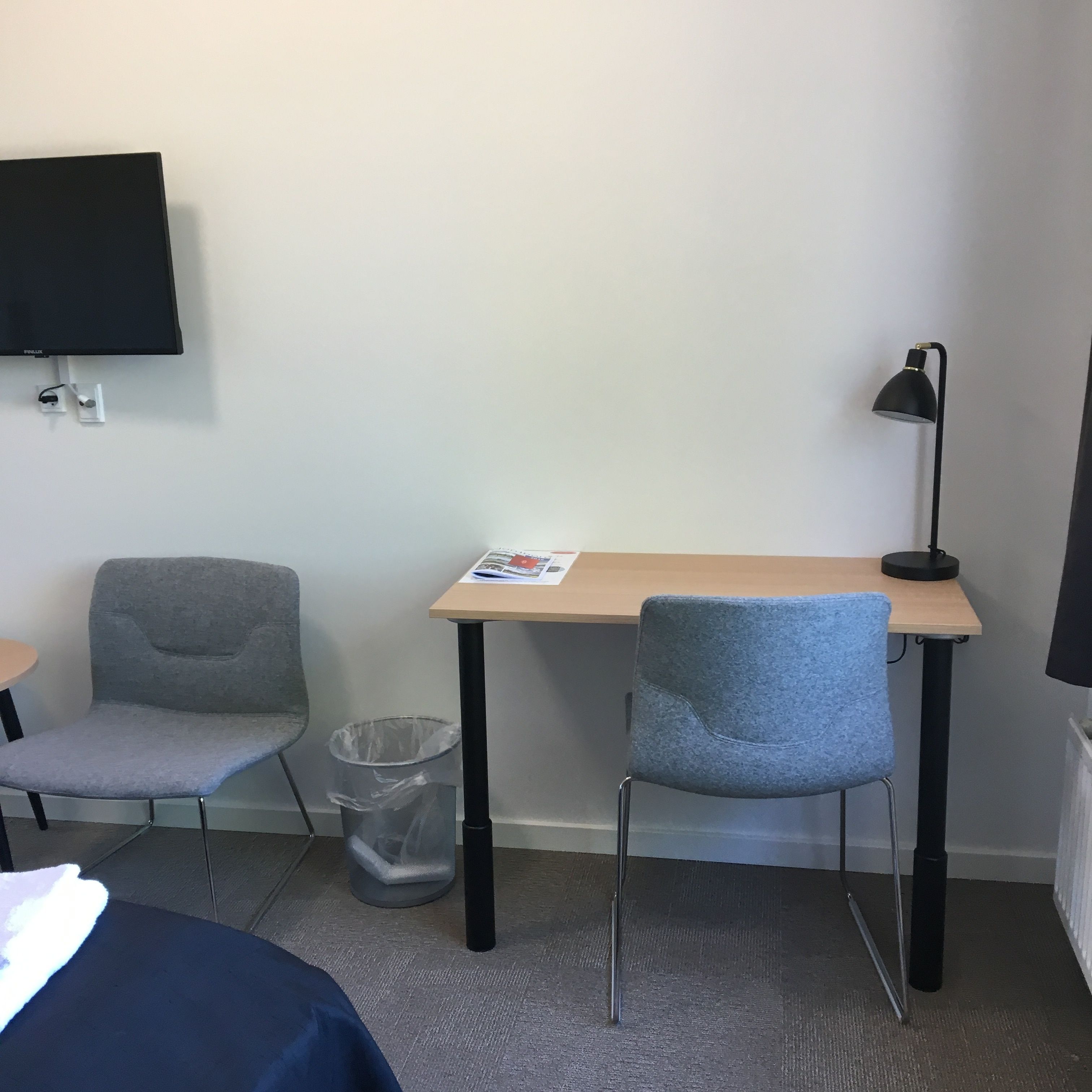 A hotel room with a bed, a desk with a chair, a TV mounted on the wall, a trash bin, and a lamp.