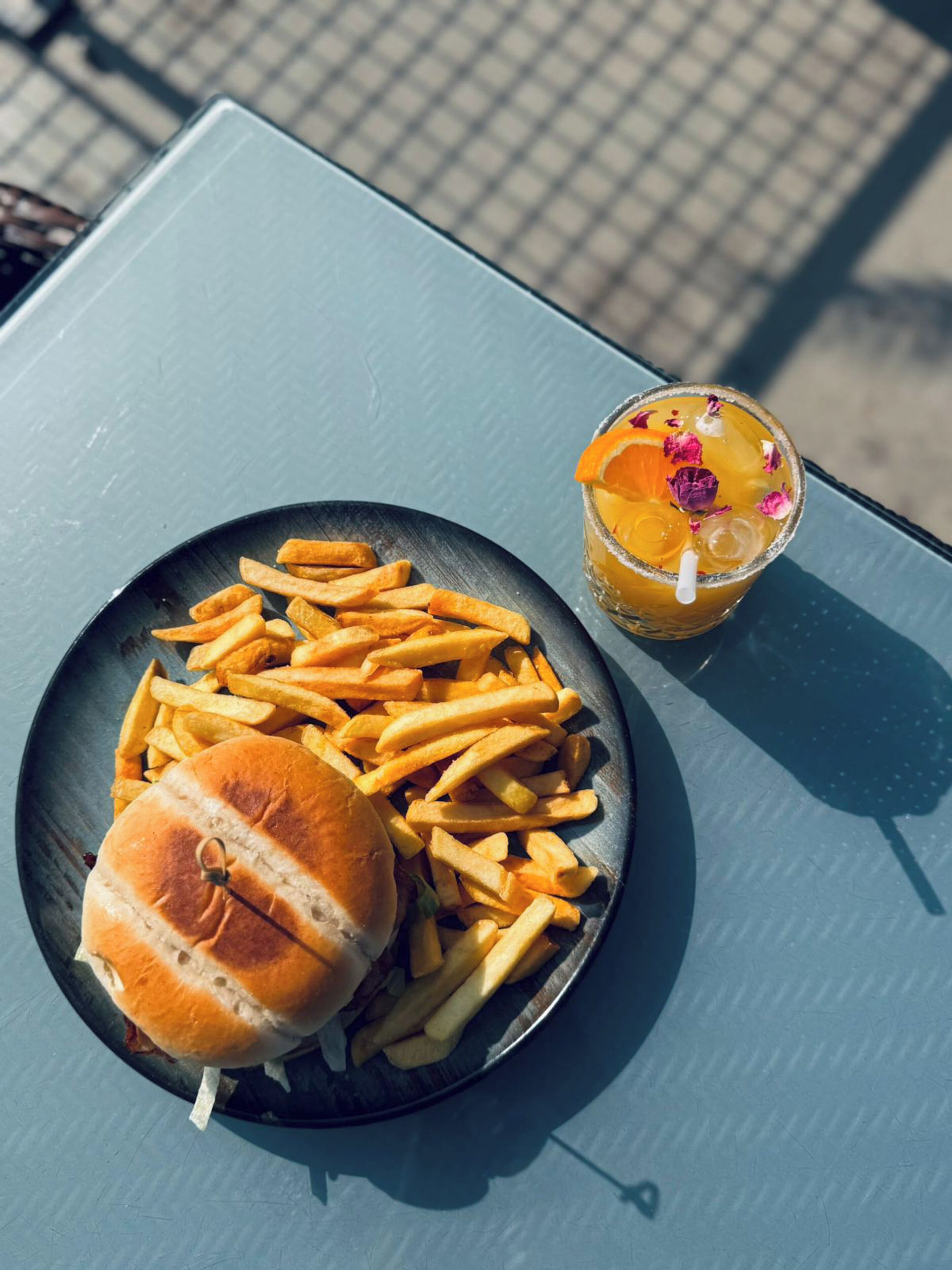 A plate of French fries and a burger on a metal tray, and a glass of iced drink with fruit garnish on a table.