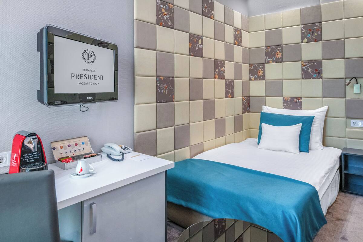A hotel room with a bed, a desk with a coffee machine, a phone, and a television displaying 'BUDAPEST PRESIDENT MOZART GROUP'.