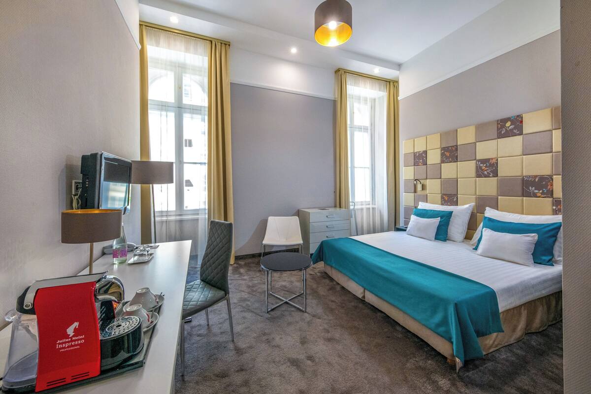 A hotel room with a bed, a desk with a coffee machine, a chair, a TV, and a window with curtains.