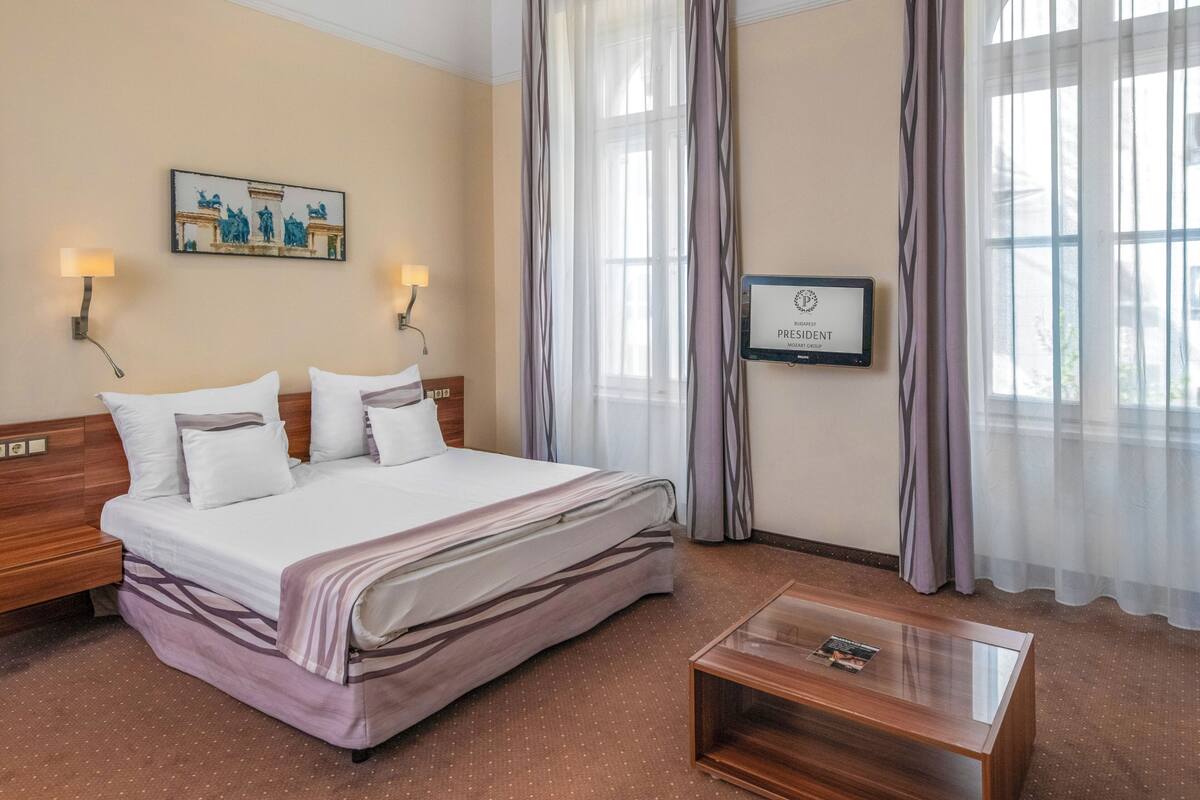 A hotel room with a bed, a nightstand, a coffee table, a wall-mounted TV, and a painting.