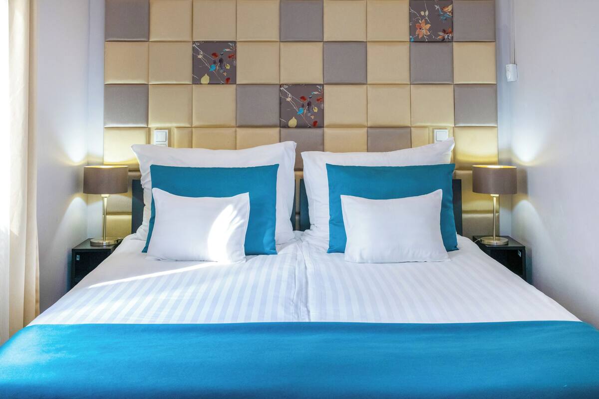 A hotel room with a large bed, two bedside tables with lamps, a headboard with a floral pattern, and a wall with a grid of square panels.