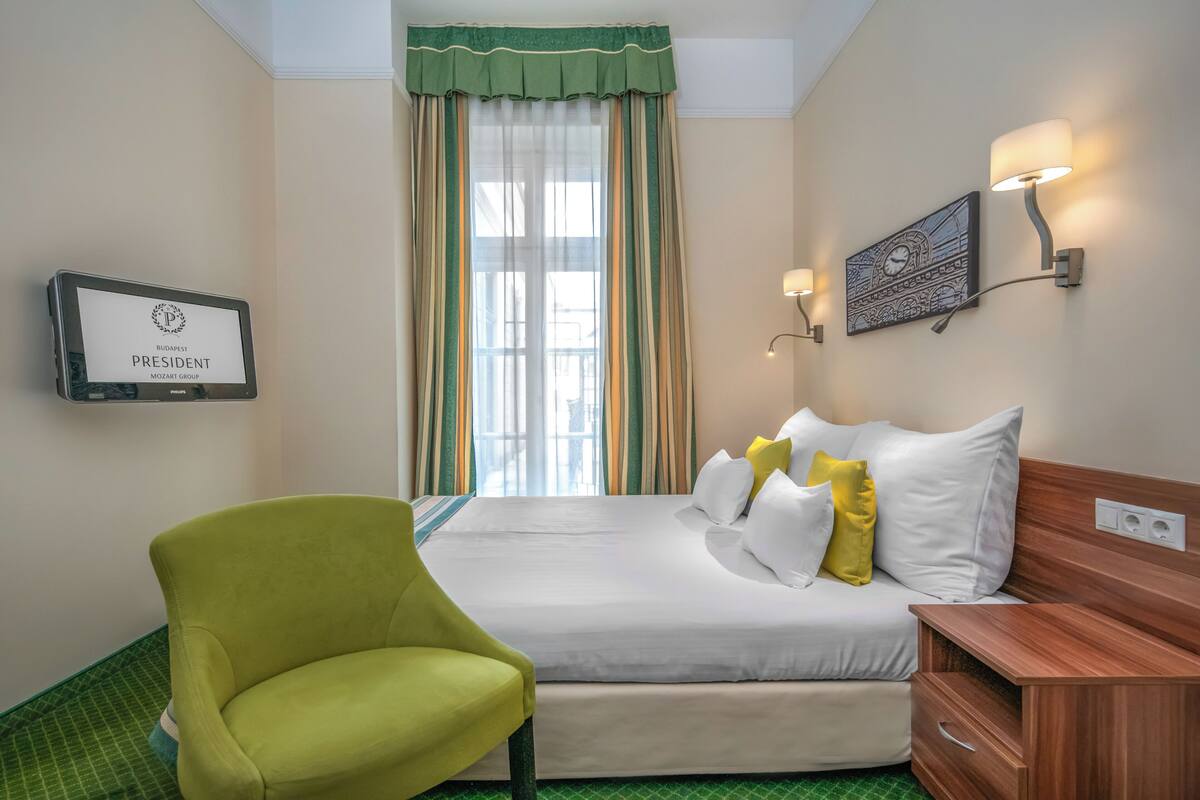 A hotel room with a bed, a green armchair, a wall-mounted TV, and a window with curtains.
