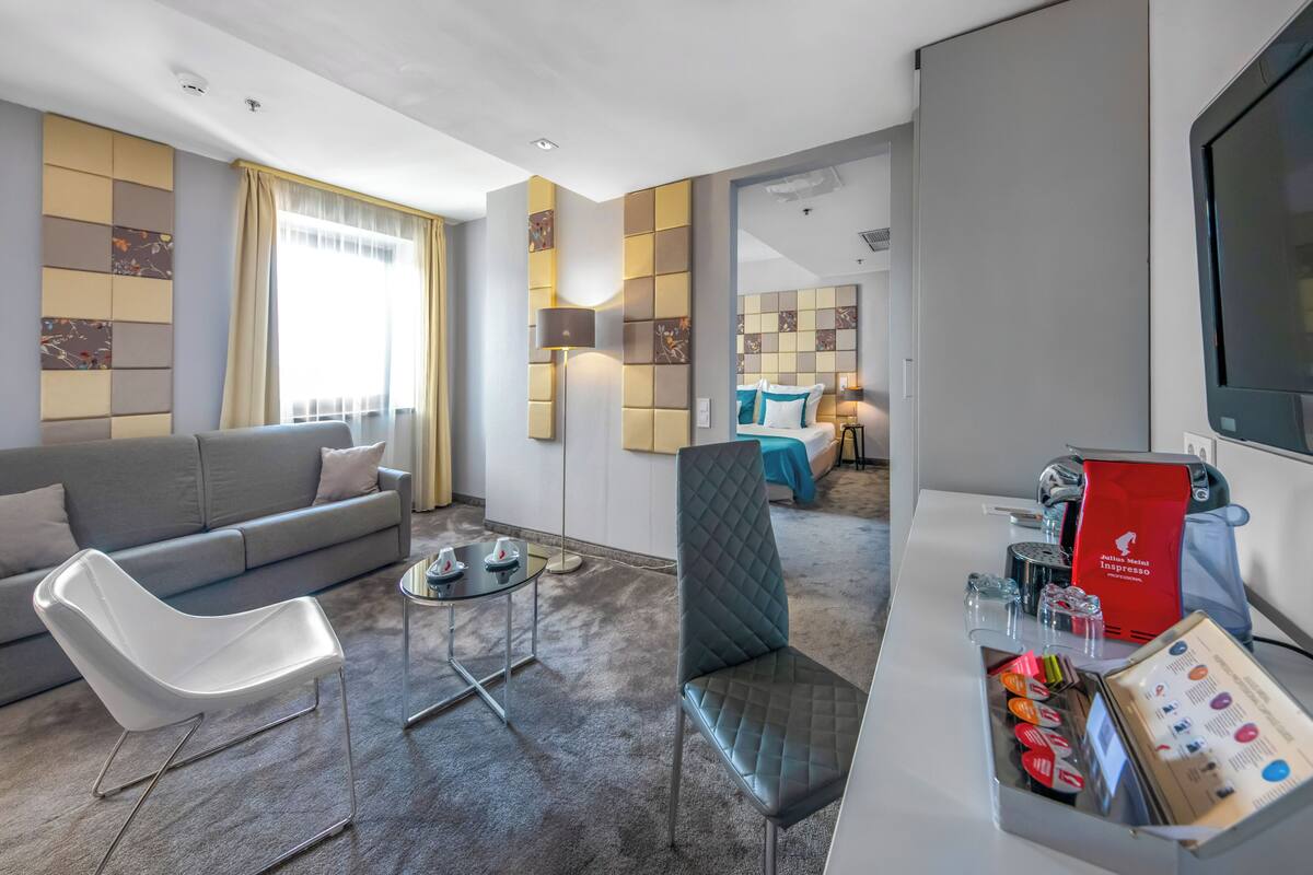 A modern hotel room with a grey sofa, a white chair, a small table, a coffee station, and a view of the adjacent room.
