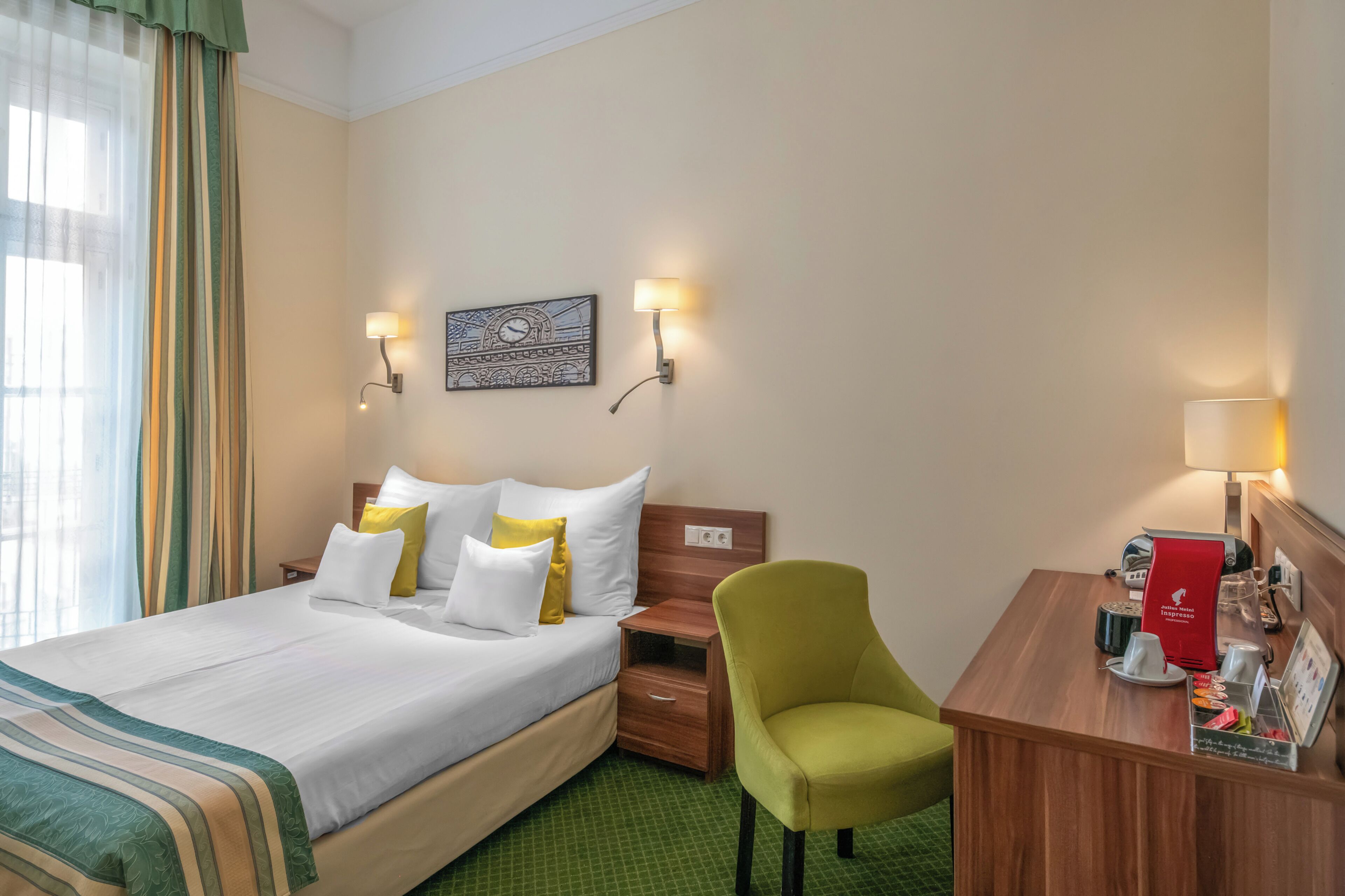 A hotel room with a bed, two bedside tables, a desk with a chair, and a wall-mounted artwork.