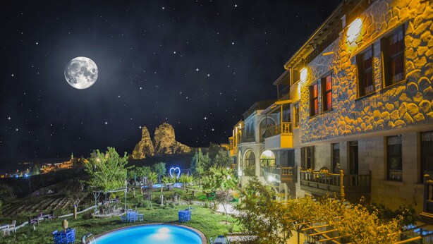 Une vue nocturne d’un hôtel avec une piscine, une architecture en pierre et une lune bien visible dans le ciel.