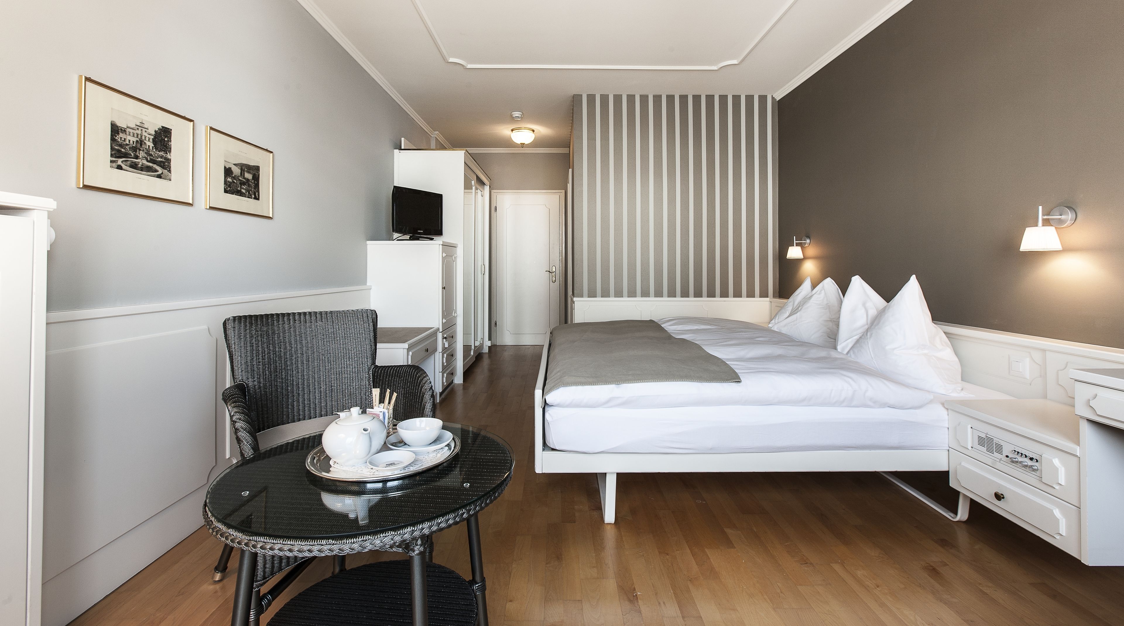 Et hotelværelse med seng, natbord, stol og et lille bord med tesæt.
