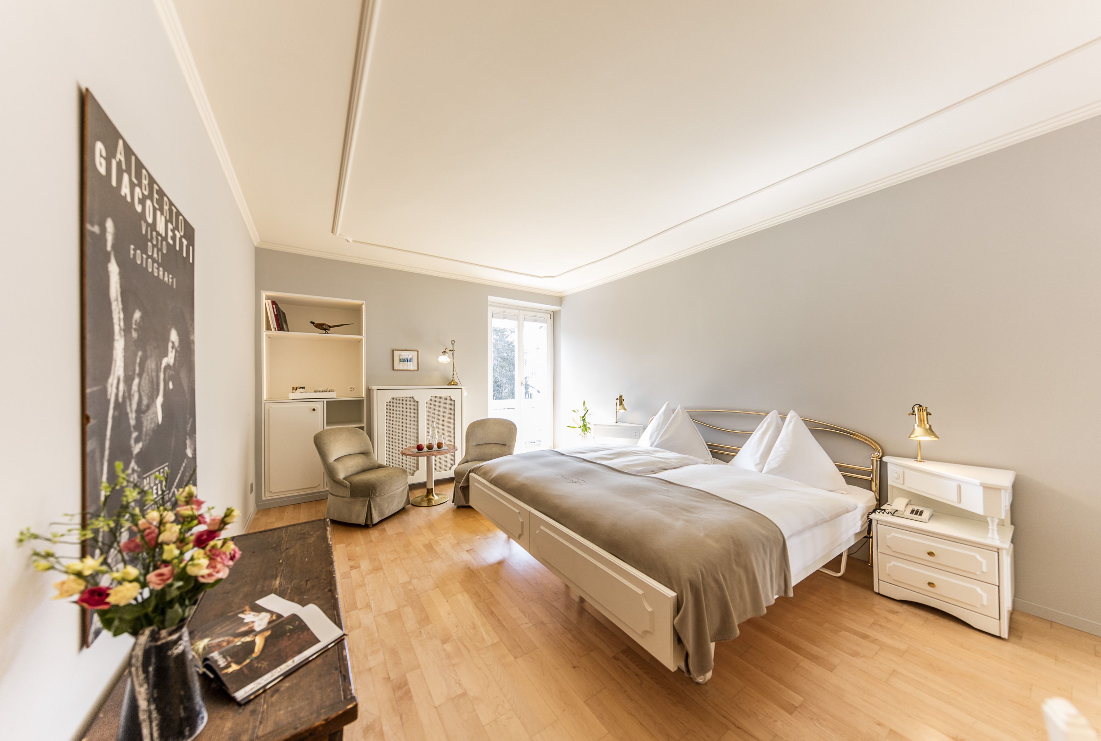 Ein Schlafzimmer mit einem Bett, einem Nachttisch, einer Lampe, einem Stuhl und einem kleinen Tisch mit Blumen.