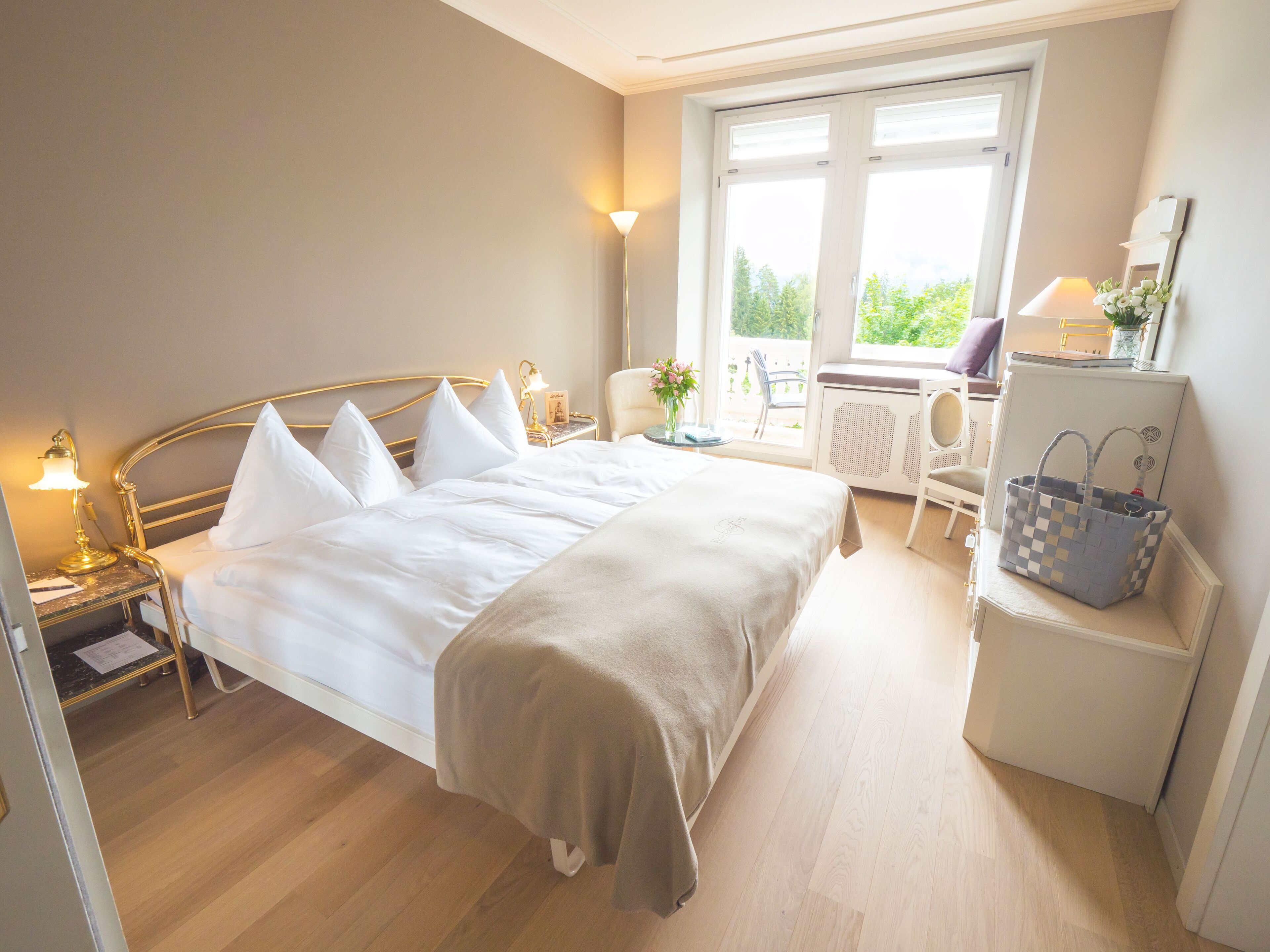 A hotel room with a bed, bedside table with a lamp, a chair, a desk with a lamp, and a window with a view of greenery.