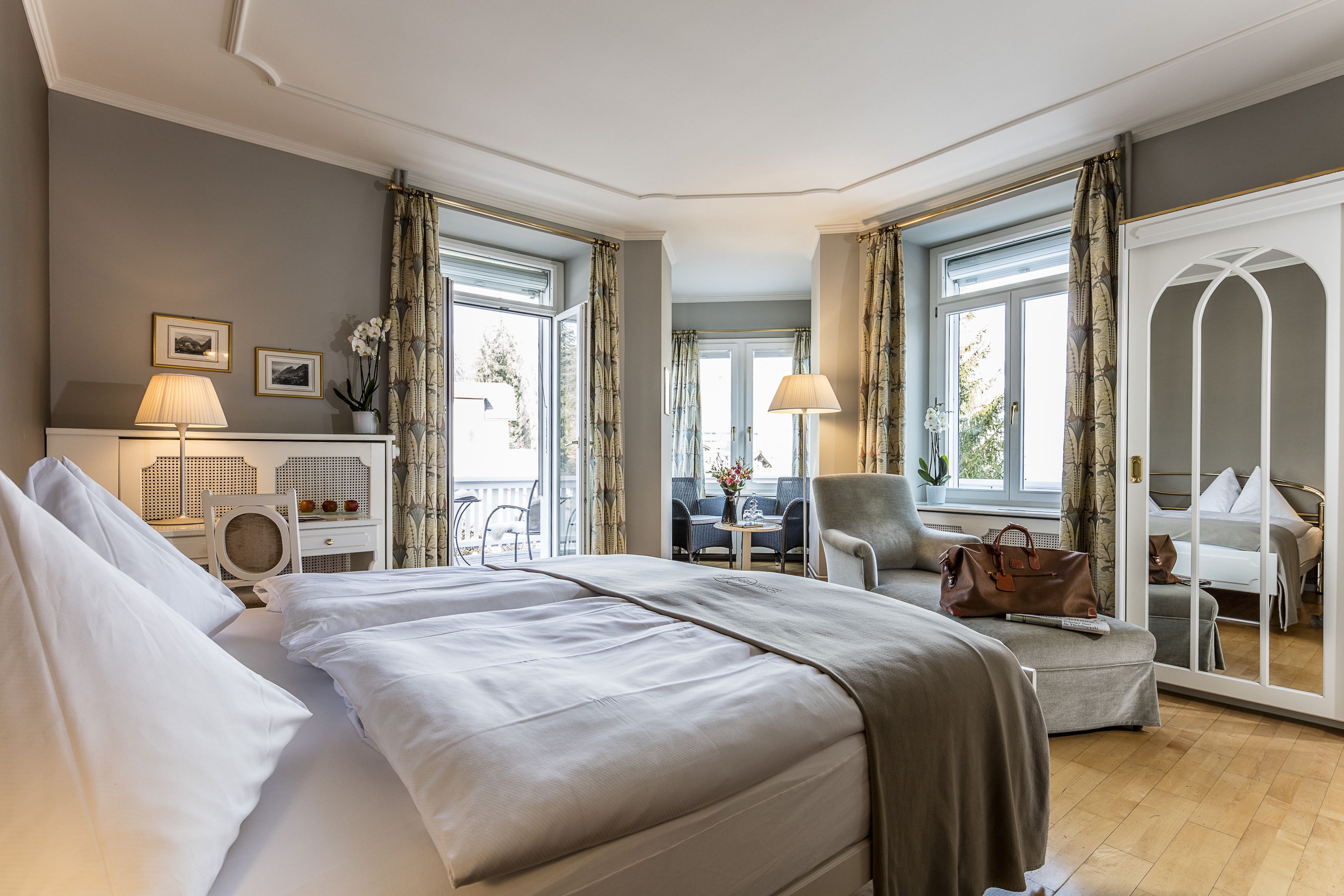 Ein Hotelzimmer mit einem großen Bett, einem Schreibtisch mit Lampe, einem Sessel und Blick ins Freie durch große Fenster.
