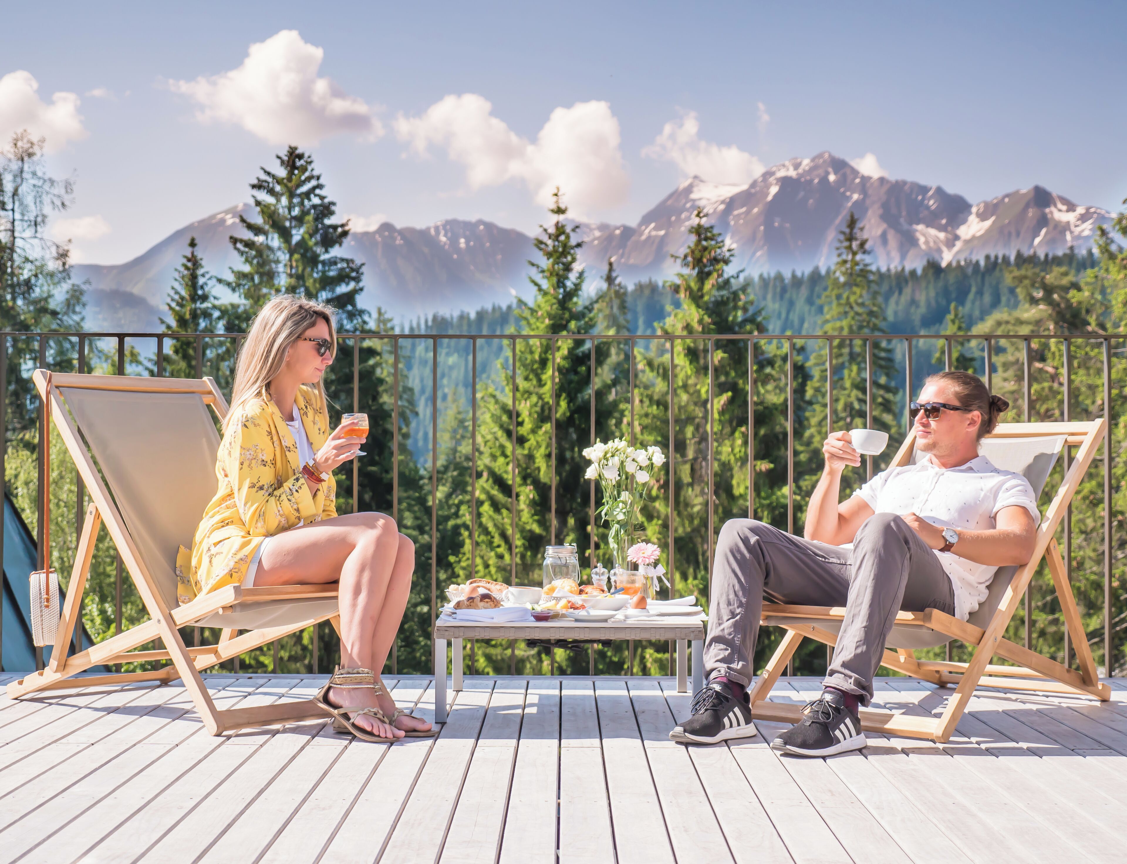 Two people seated on deck chairs with a table between them, overlooking a mountainous landscape.