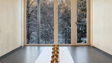 Una sala de meditación con interior de madera, un ventanal con vistas a árboles cubiertos de nieve y una fila de cuencos dorados sobre un tapete blanco.