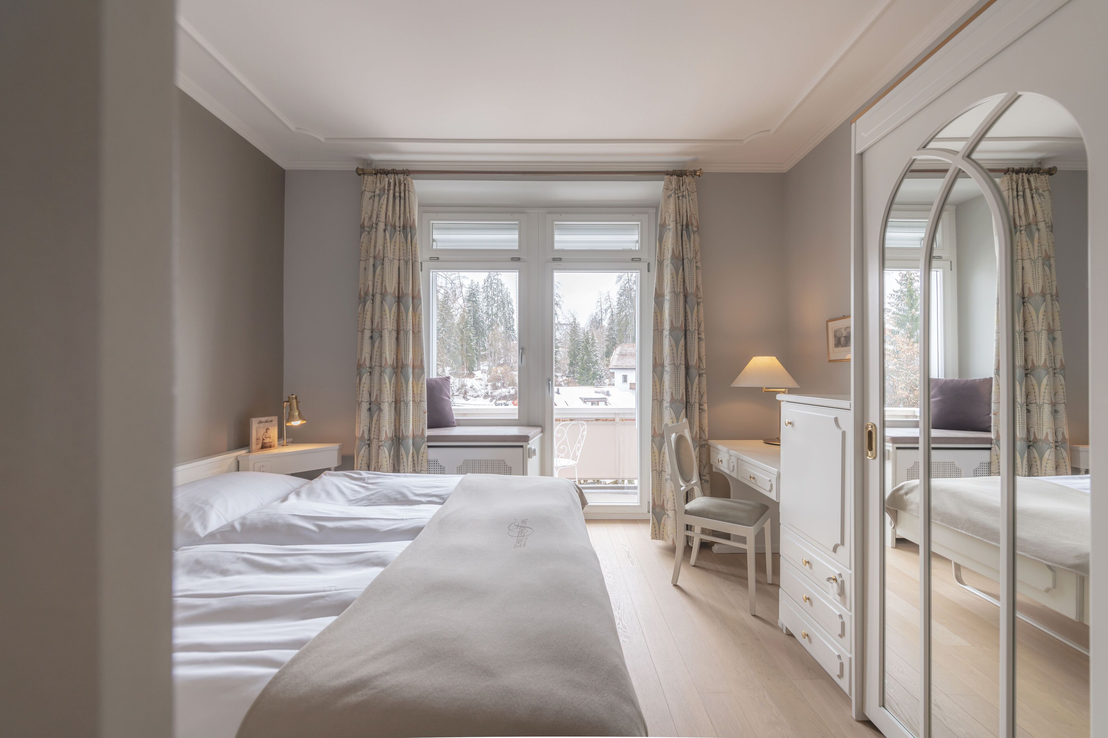 Ein Hotelzimmer mit einem großen Bett, einem Schreibtisch, einem Sessel, einem Schminktisch und einem großen Spiegel. Zwei Fenster mit Vorhängen bieten Blick auf Bäume und Gebäude.