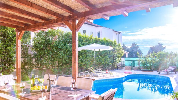 An outdoor pool area with a wooden pergola, lounge chairs, and a dining table set for a meal. - Villa Mare (Hvar)