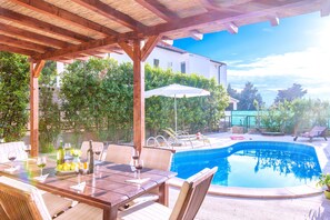 An outdoor pool area with a wooden pergola, lounge chairs, and a dining table set for a meal. - Villa Mare (Hvar)
