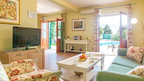 A living room with a television, sofa, floral chairs, a dining table with fruit, and a view of a swimming pool. - Villa Mare (Hvar)
