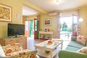 A living room with a television, sofa, floral chairs, a dining table with fruit, and a view of a swimming pool. - Villa Mare (Hvar)