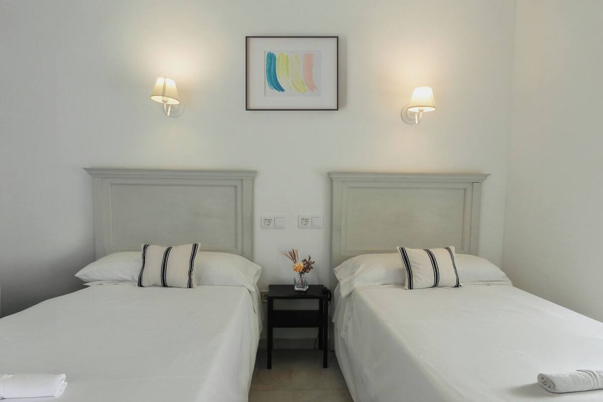 A hotel room with two single beds, each with a pillow and a small bedside table with a lamp.