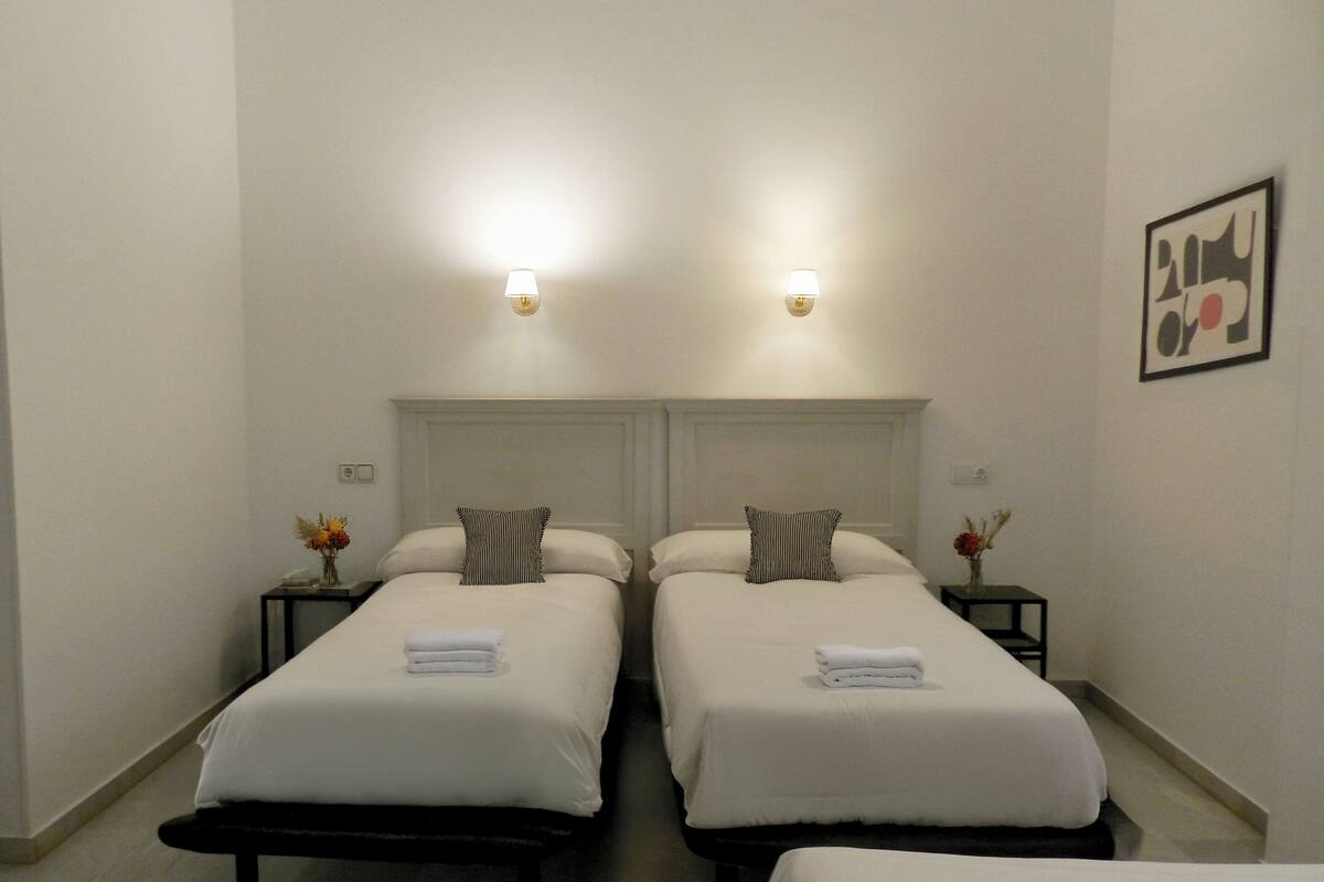 A hotel room with two single beds, each with a pillow and a folded towel. There are bedside tables with lamps and a vase of flowers.