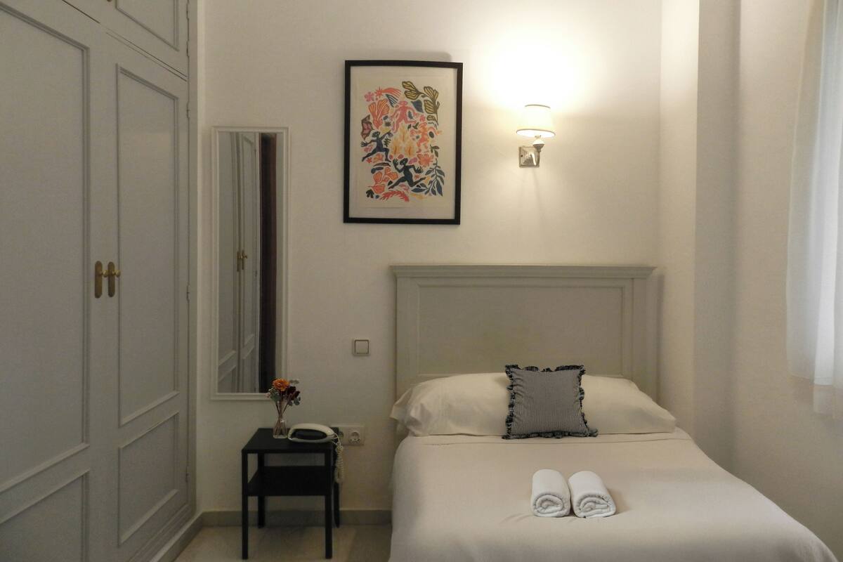 A hotel room with a bed, a bedside table with a phone, a framed artwork, and a wall-mounted light.