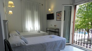 A hotel room with a bed, a desk, a chair, a TV, and a balcony with a view of trees. - Hotel Doña Blanca (Seville)