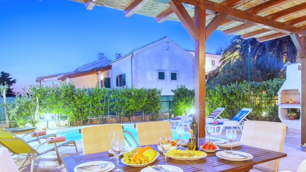 A dining area with a table set for a meal, outdoor chairs, and a pool in the background. - Villa Cvita (Hvar)