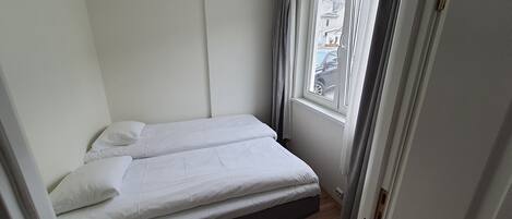 Una habitación pequeña con una cama individual, ventana con cortinas, puerta y piso de madera.