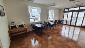 A dining area with a long table, chairs, a microwave, and windows with curtains. - Systra Hotel Søvik (Ålesund)
