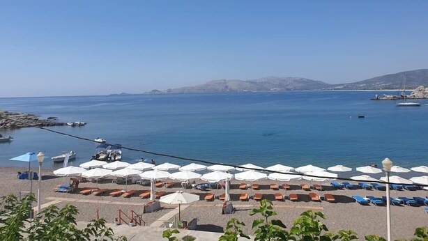 Vista de la playa con sombrillas y sillas de descanso, barcos en el agua y cielo despejado.