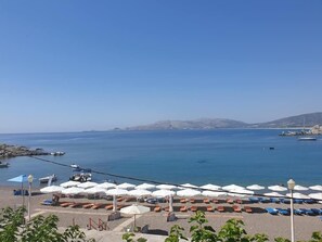 Vista de la playa con sombrillas y sillas de descanso, barcos en el agua y cielo despejado.