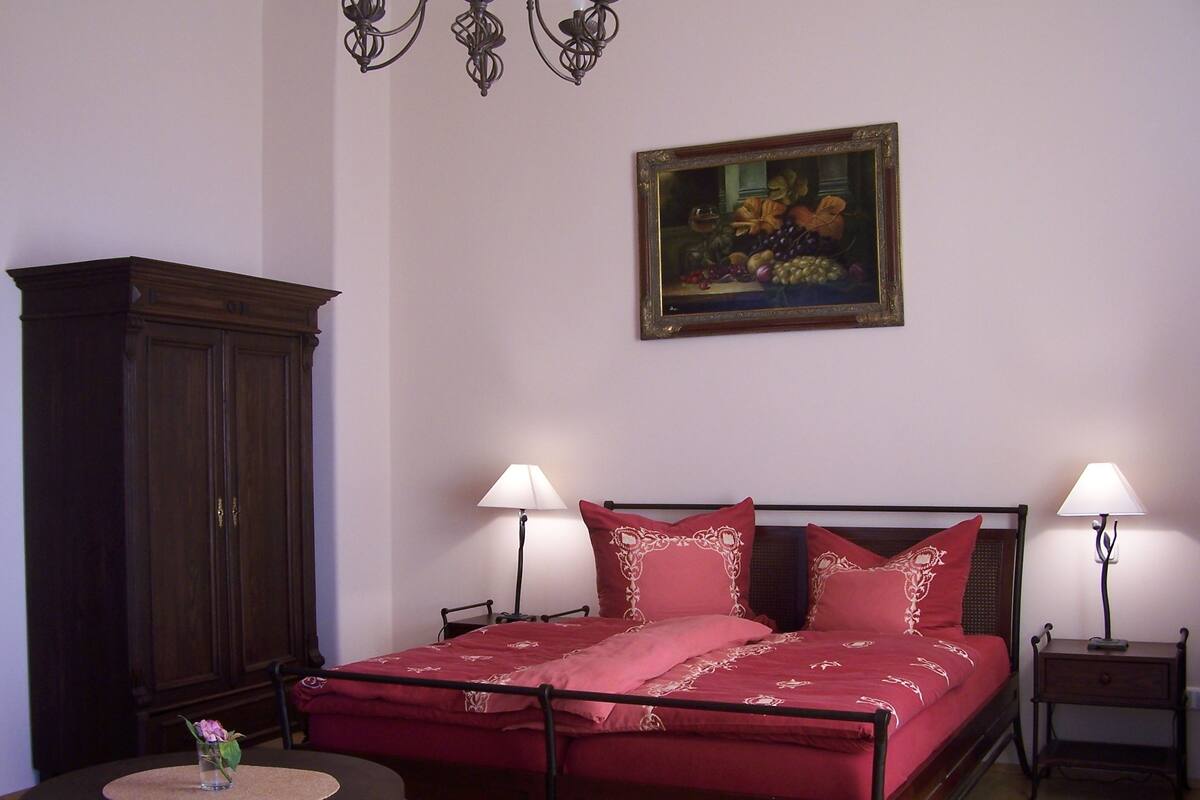 A bedroom with a bed, two bedside lamps, a wooden cabinet, a round table with a vase, and a painting on the wall.