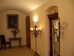 A hallway with a wooden chair, a table with a vase of flowers, a painting on the wall, and a mirror with a decorative stand.
