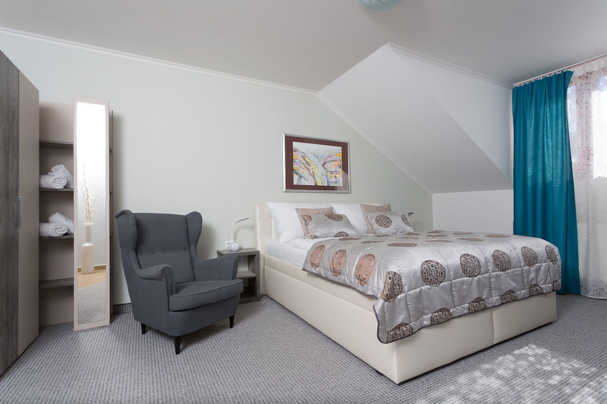 Un dormitorio con cama, una silla, una mesita de noche, un espejo y una ventana con cortinas.