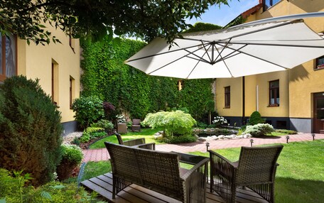 A patio area with wicker furniture, a large umbrella, and a garden with hedges and a small tree.