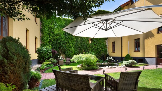 Un patio con mobiliario de mimbre, un paraguas grande y un jardín con setos y un árbol pequeño.