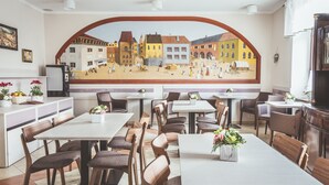 Un restaurante con un mural que representa una escena urbana, mesas y sillas de madera, y una barra blanca con un refrigerador.