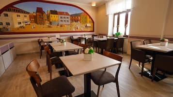 A restaurant with a mural of a village scene on the wall, wooden tables and chairs, and potted plants on the windowsills.