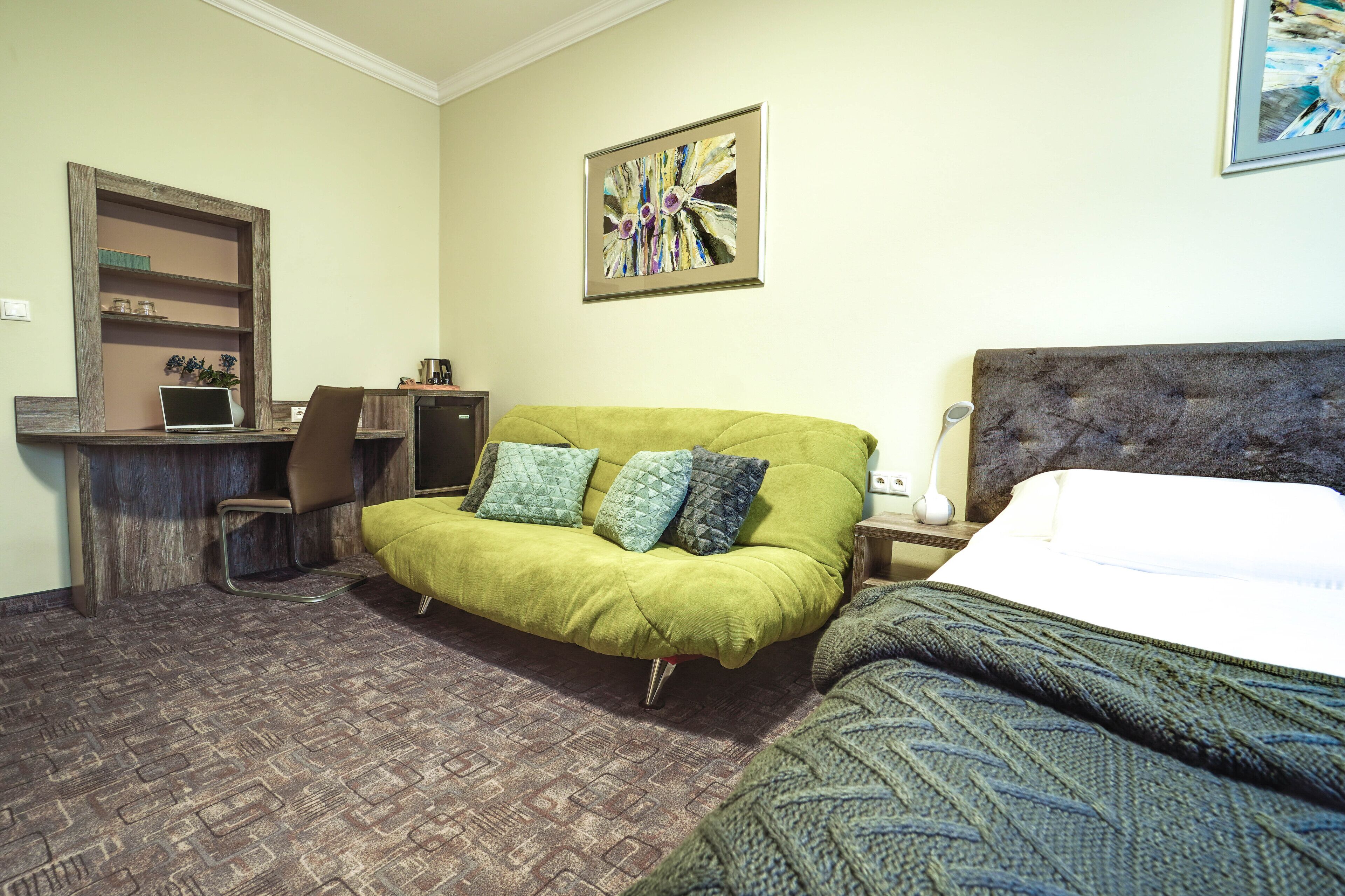 Une chambre d’hôtel avec un canapé vert, un lit recouvert d’une couette blanche, un bureau avec un ordinateur portable et un tableau abstrait encadré accroché au mur.